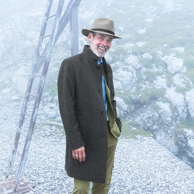 Älterer Mann mit grauem Hut und dunklem Mantel steht lächelnd in einer nebligen Berglandschaft – stilvolle Herbst- und Wintermode für Herren.