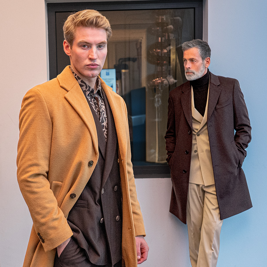 Zwei Männer in eleganten Mänteln – einer im hellbraunen Mantel mit dunklem Anzug, der andere im dunkelbraunen Mantel mit hellem Anzug – moderne Businessmode im Stil des stillen Luxus.