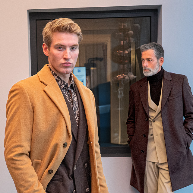Zwei Männer in eleganten Mänteln – einer im hellbraunen Mantel mit dunklem Anzug, der andere im dunkelbraunen Mantel mit hellem Anzug – moderne Businessmode im Stil des stillen Luxus.