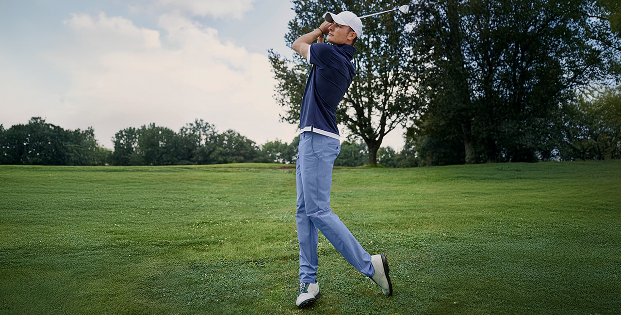 Ein junger Mann in sportlicher Golfbekleidung schlägt einen Golfschläger auf einem gepflegten Golfplatz. Er trägt ein dunkelblaues Poloshirt mit weißen Akzenten, eine weiße Baseballkappe und hellblaue Hosen.