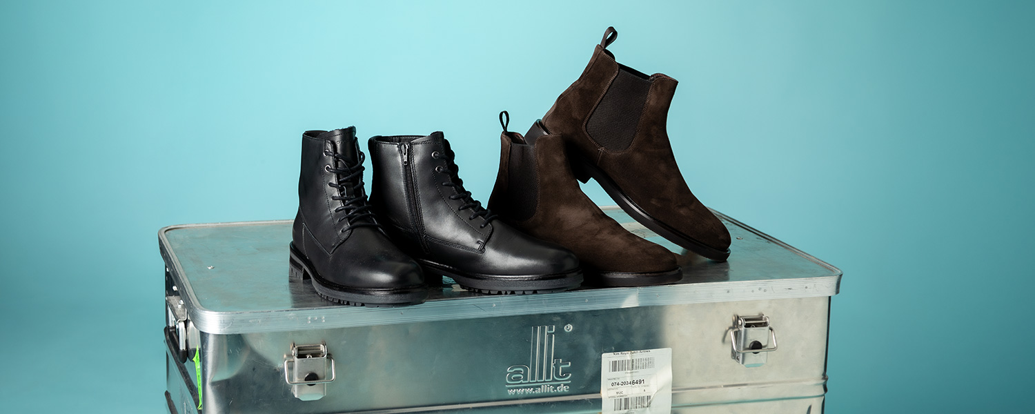 Schwarze Lederstiefel und braune Wildleder-Chelsea-Boots auf Metallkiste vor hellblauem Hintergrund.
