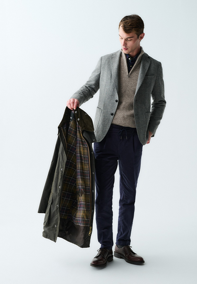 Herr in grauem Blazer und beige Strickpullover, navy Chinos, hält Jacke mit kariertem Muster