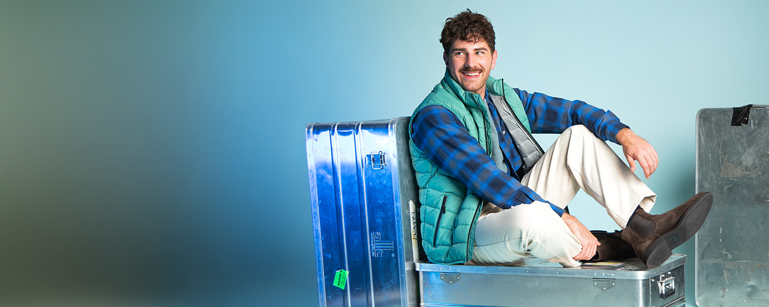 Mann in blau-grün karierter Jacke mit grüner Steppweste auf Metallkisten sitzend, lässige Kleidung