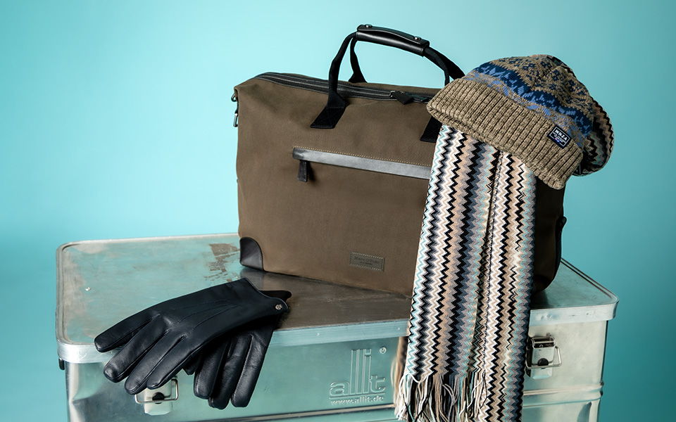 Braune Reisetasche mit Lederdetails, schwarze Lederhandschuhe, gemusterter Schal und Strickmütze auf Metallkoffer platziert
