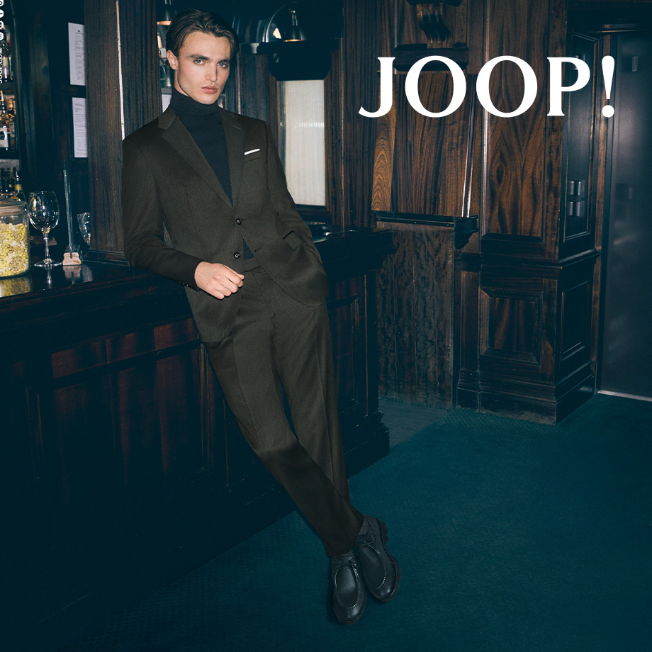 Mann in dunkelbraunem Anzug mit schwarzem Rollkragenpullover, lässig an Bar gelehnt