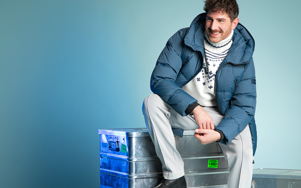 Mann in blauer Daunenjacke und Winterpullover, lässig sitzend auf Metallkiste