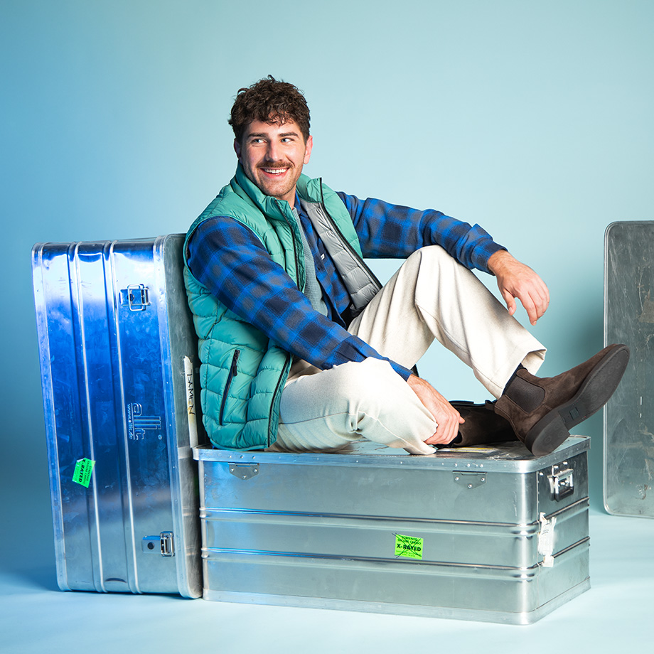 Mann in karierter Wolljacke mit grüner Steppweste und beigener Hose sitzend, legere Modekombination