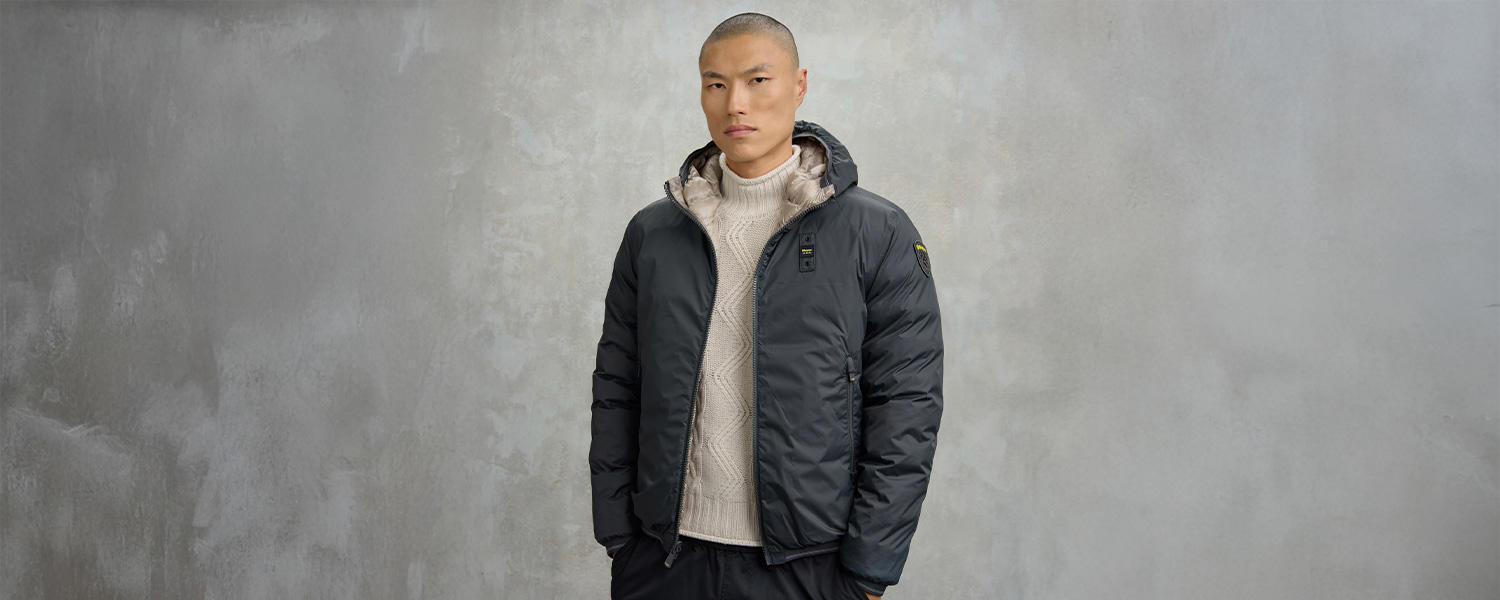 Mann in dunkelgrauer Steppjacke mit Kapuze und beigem Strickpullover vor neutralem Hintergrund