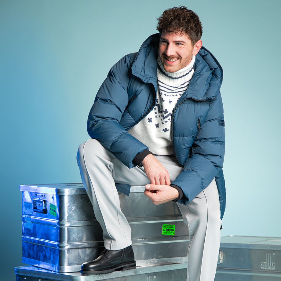 Mann in blauer Daunenjacke und Winterpullover, lässig sitzend auf Metallkiste