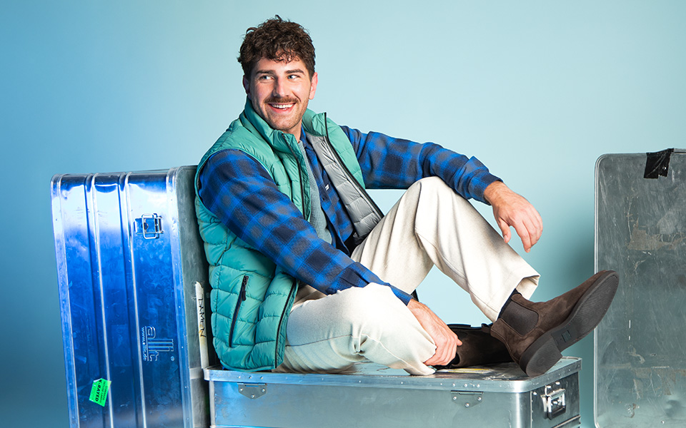 Mann in karierter Wolljacke mit grüner Steppweste und beigener Hose sitzend, legere Modekombination