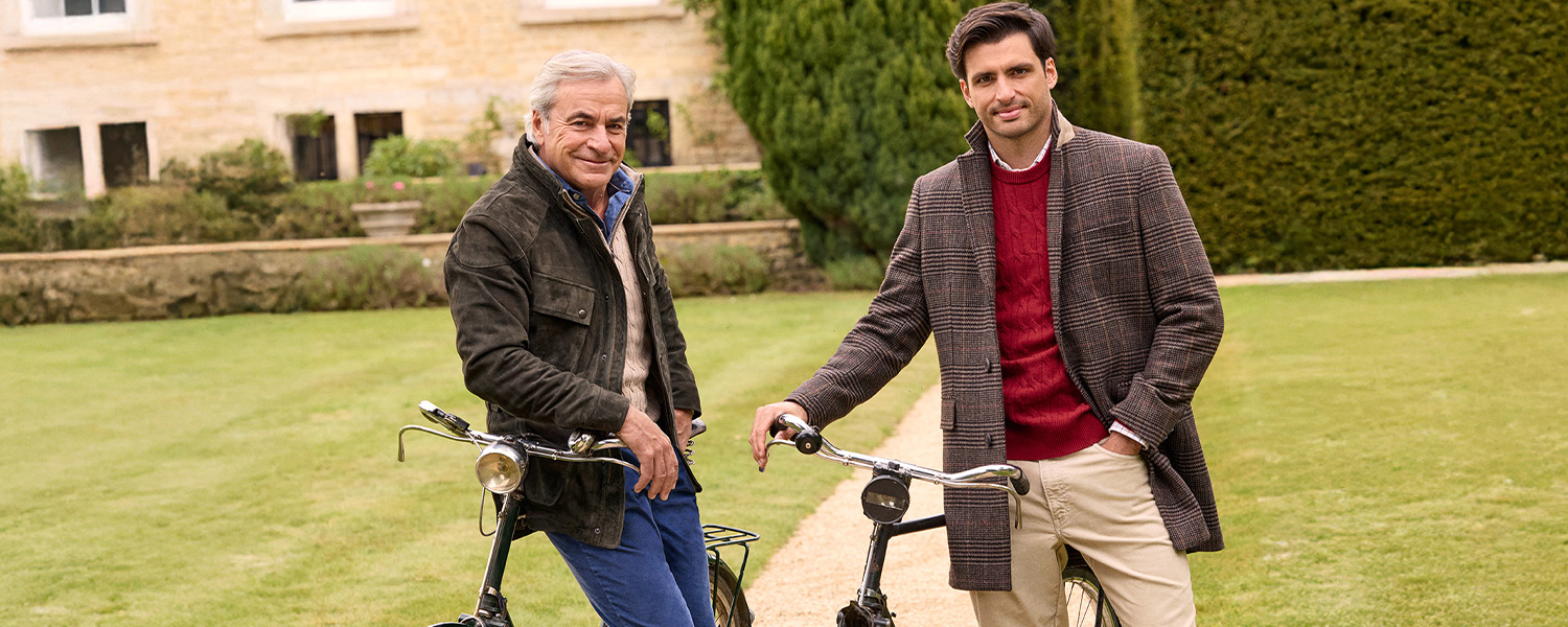 Zwei Männer in karierten Mänteln mit Rollkragenpullover und Oxford-Hemd im Garten, an Fahrräder gelehnt.