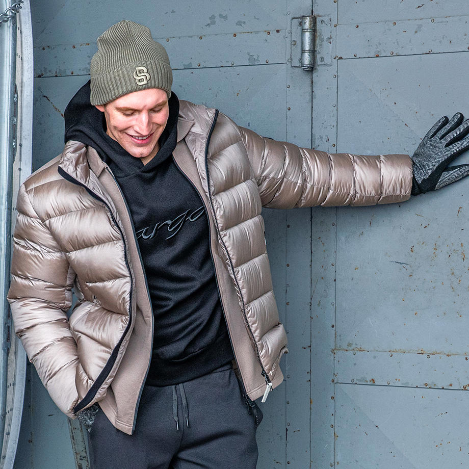 Mann in grauer Daunenjacke, schwarzem Hoodie und Beanie lässig angelehnt
