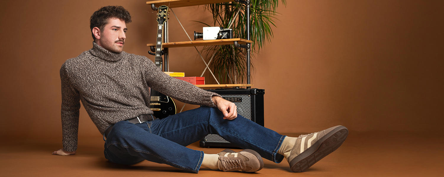 Mann in grauem Strickpullover und blauer Jeans, sitzt lässig auf braunem Hintergrund