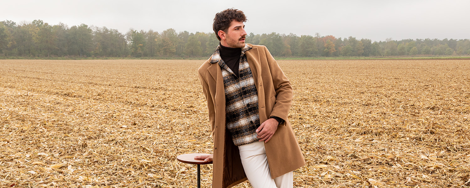Mann in kariertem Pullover mit beigem Mantel und weißen Hosen, lässig im Freien