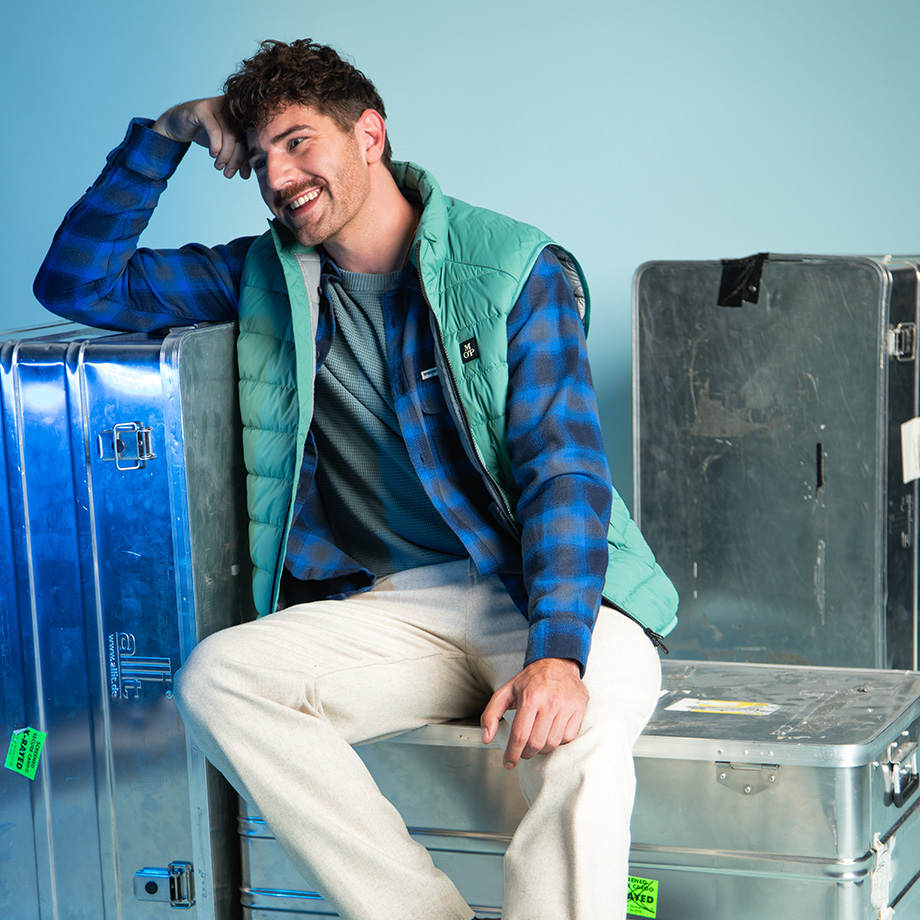 Mann in grüner Steppweste und blau kariertem Hemd, auf Metallkoffer sitzend