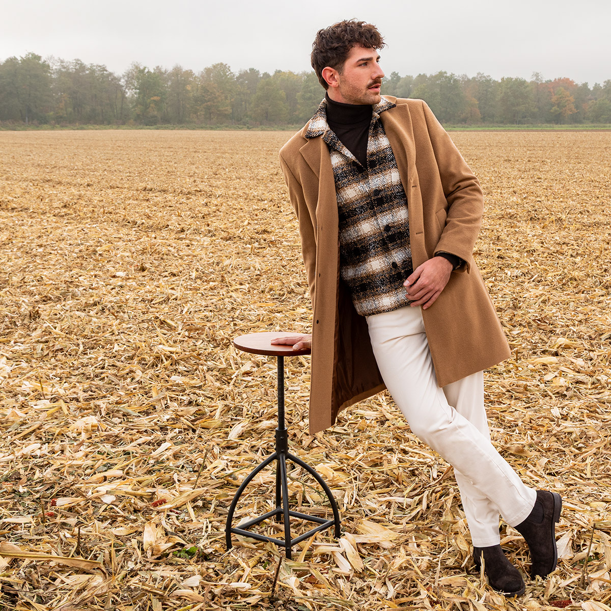 Mann in kariertem Pullover, braunem Mantel und weißen Hosen auf Feld, lässig stehend