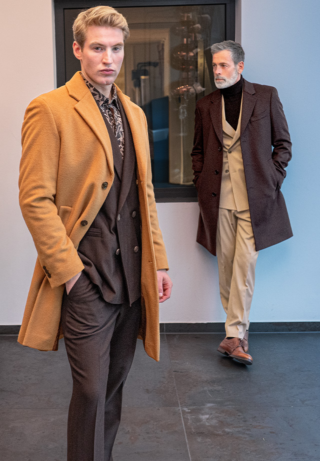 Männer in eleganten Wintermänteln, einer in camel, einer in dunkelbraun, modern kombiniert im Innenraum