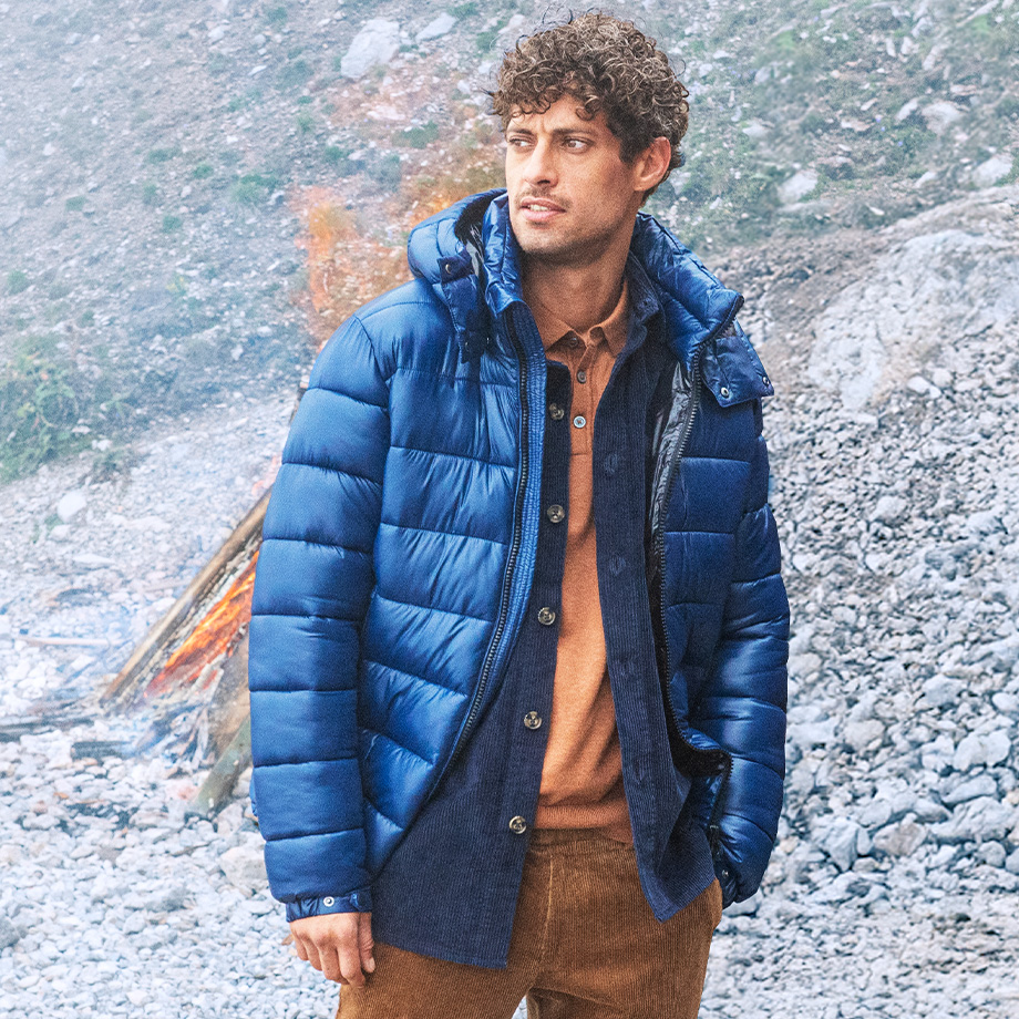 Mann in blauer Daunenjacke, braune Hose, vor Lagerfeuer, Outdoor-Look