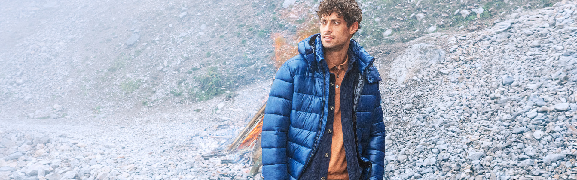 Mann in blauer Daunenjacke, braune Hose, vor Lagerfeuer, Outdoor-Look