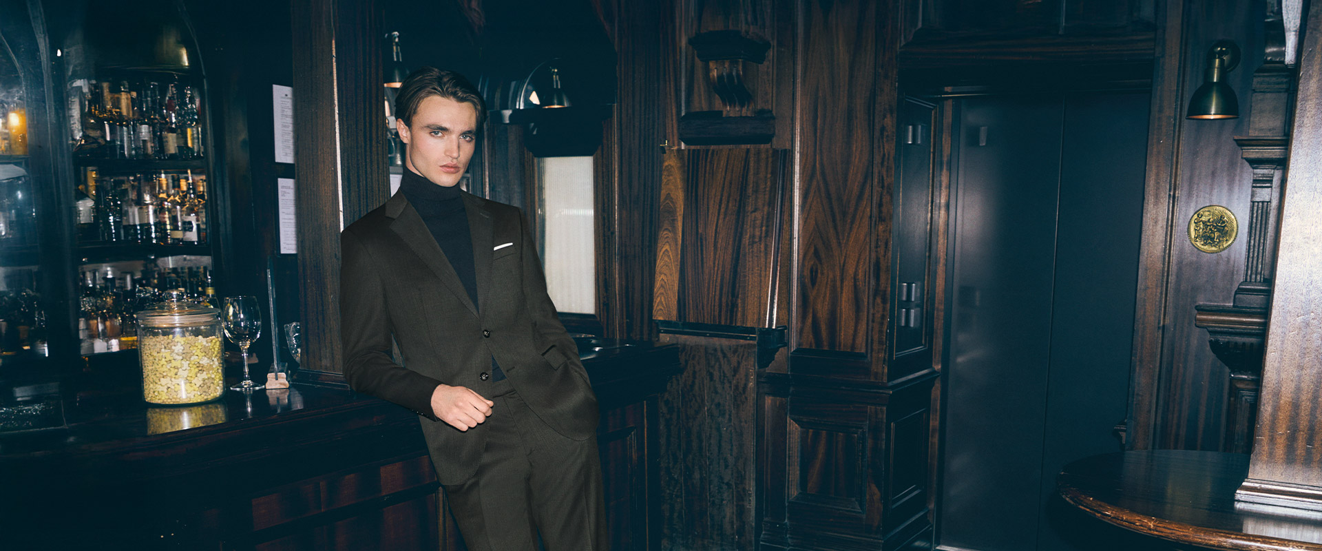 Mann in dunkelbraunem Anzug mit schwarzem Rollkragenpullover, lässig an Bar gelehnt