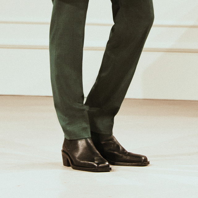 Schwarze Lederschuhe zu dunkelgrüner Stoffhose auf hellem Boden