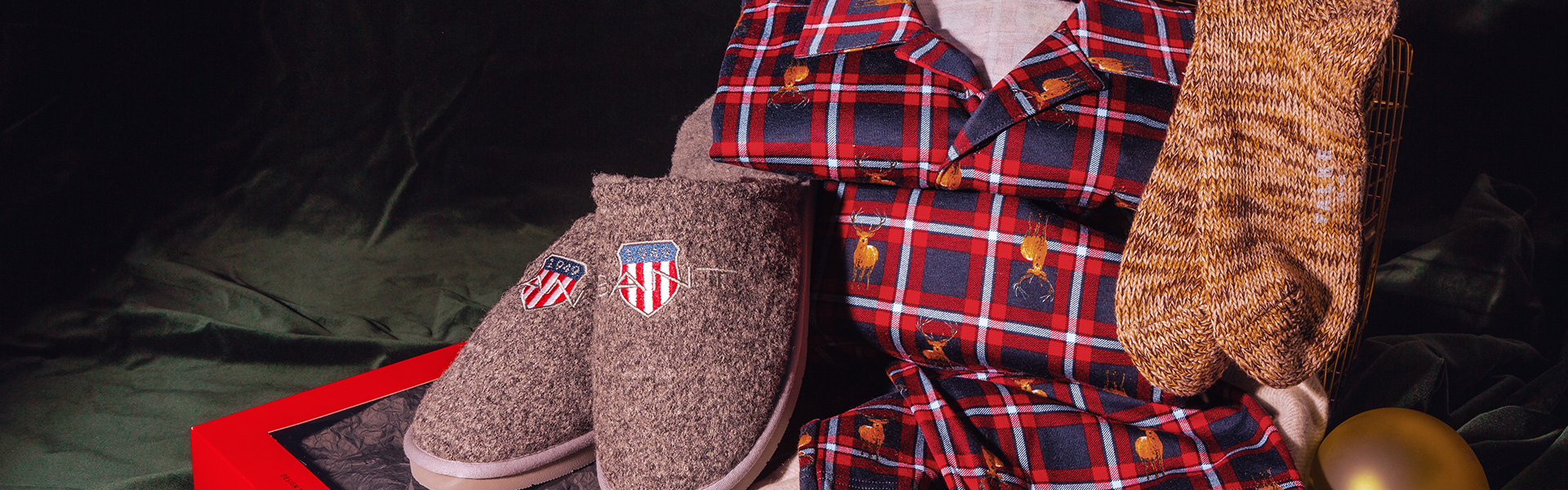 Rotes kariertes Pyjama-Set mit Hirschmuster auf Wollschal, graue Filzpantoffeln und braune Stricksocken.