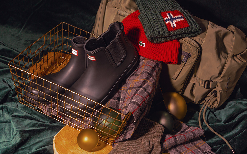 Schwarze Gummistiefel in Drahtkorb, mit rotem Wollschal und beigem Rucksack drapiert