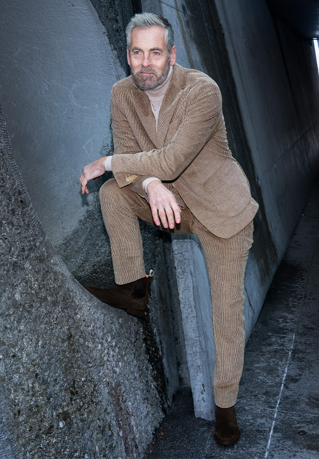Mann in beigefarbenem Cordanzug mit Rollkragenpullover, angelehnt an Betonwand