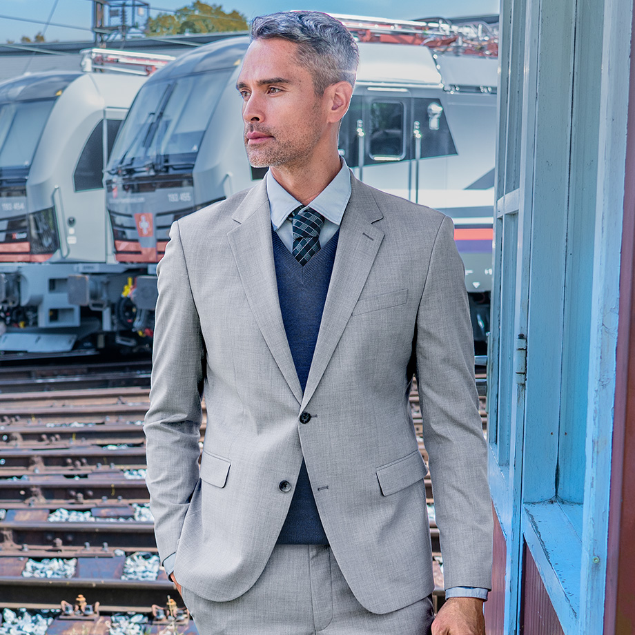 Hellgrauer Anzug aus Wolle mit dunkelblauem V-Ausschnitt-Pullover, getragen von Mann an Bahngleisen