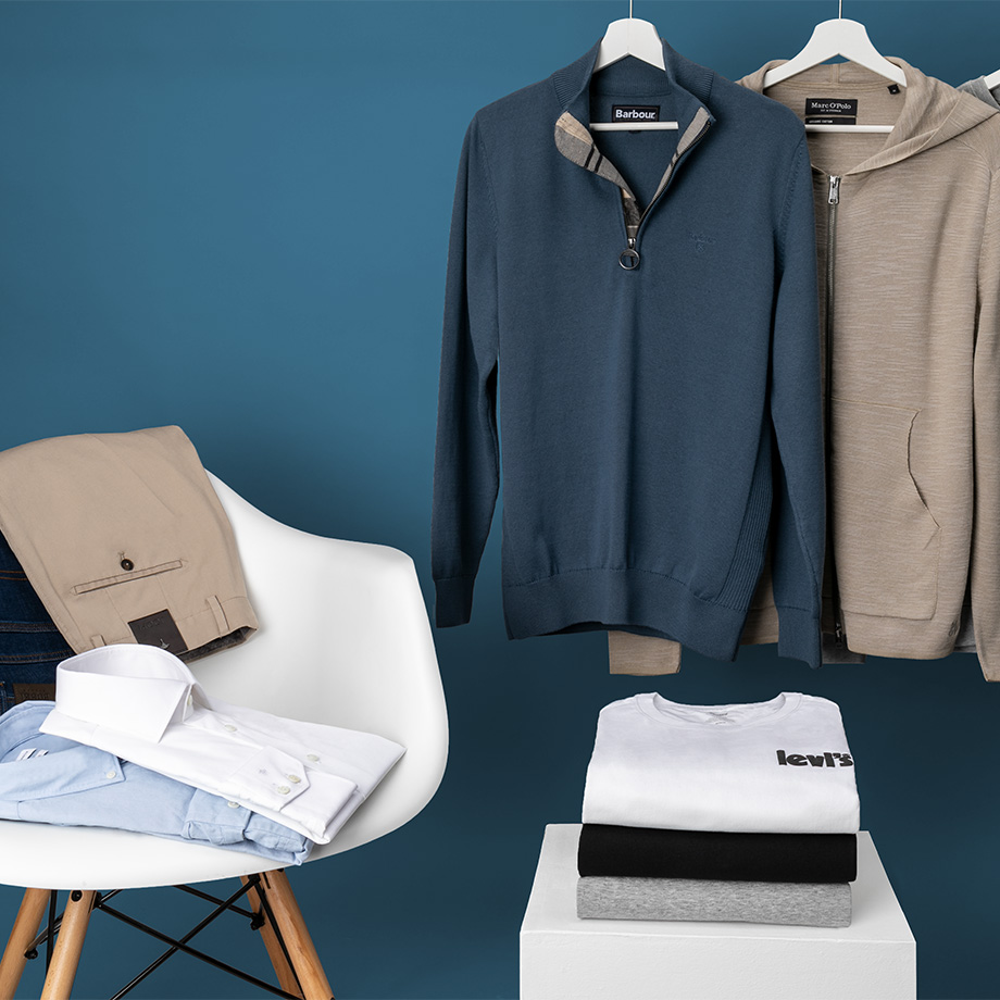 Beigefarbene Chinohose und weißes Business-Hemd auf Stuhl, blauer Pullover und grauer Hoodie hängend, gefaltete T-Shirts.