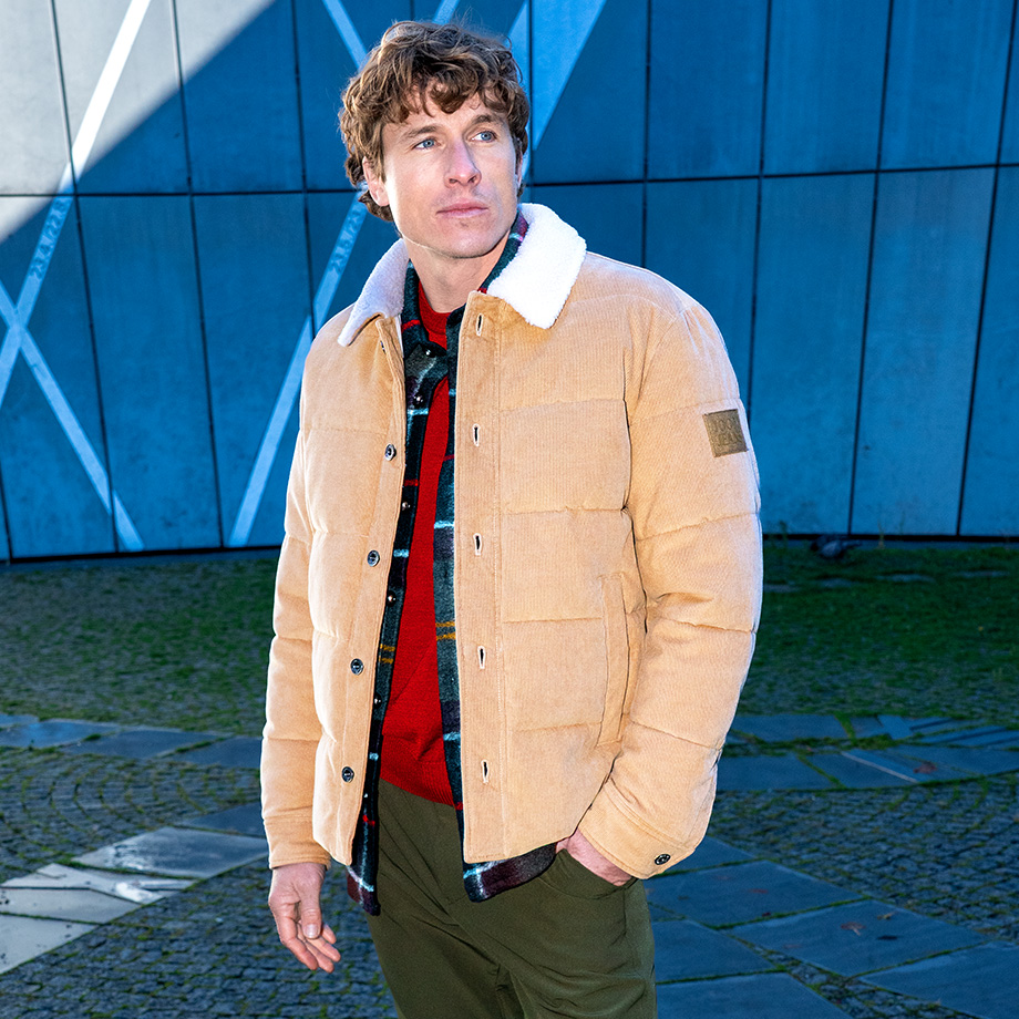 Mann in beiger Steppjacke mit weißem Kragen, roter Pullover, grüne Hose, weiße Sneaker