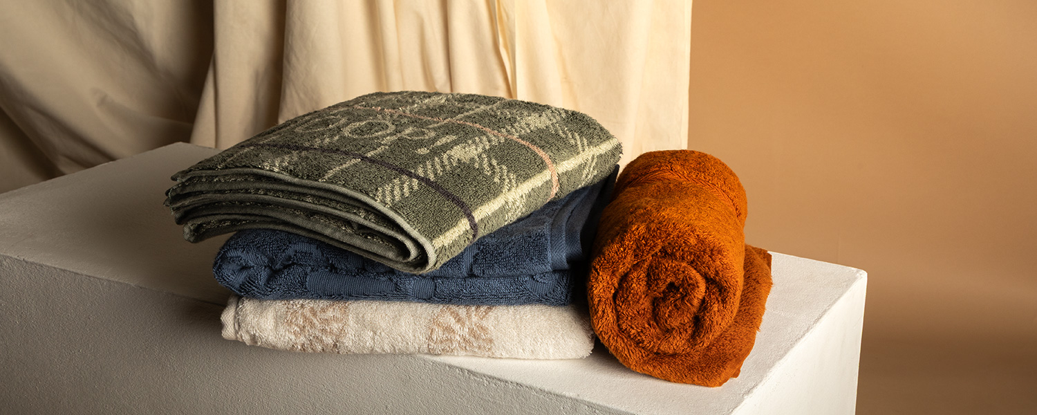 Gestapelte Handtücher in olivgrünem Karo, marineblau, beige und rostbraun aus Baumwolle auf weißem Podest.