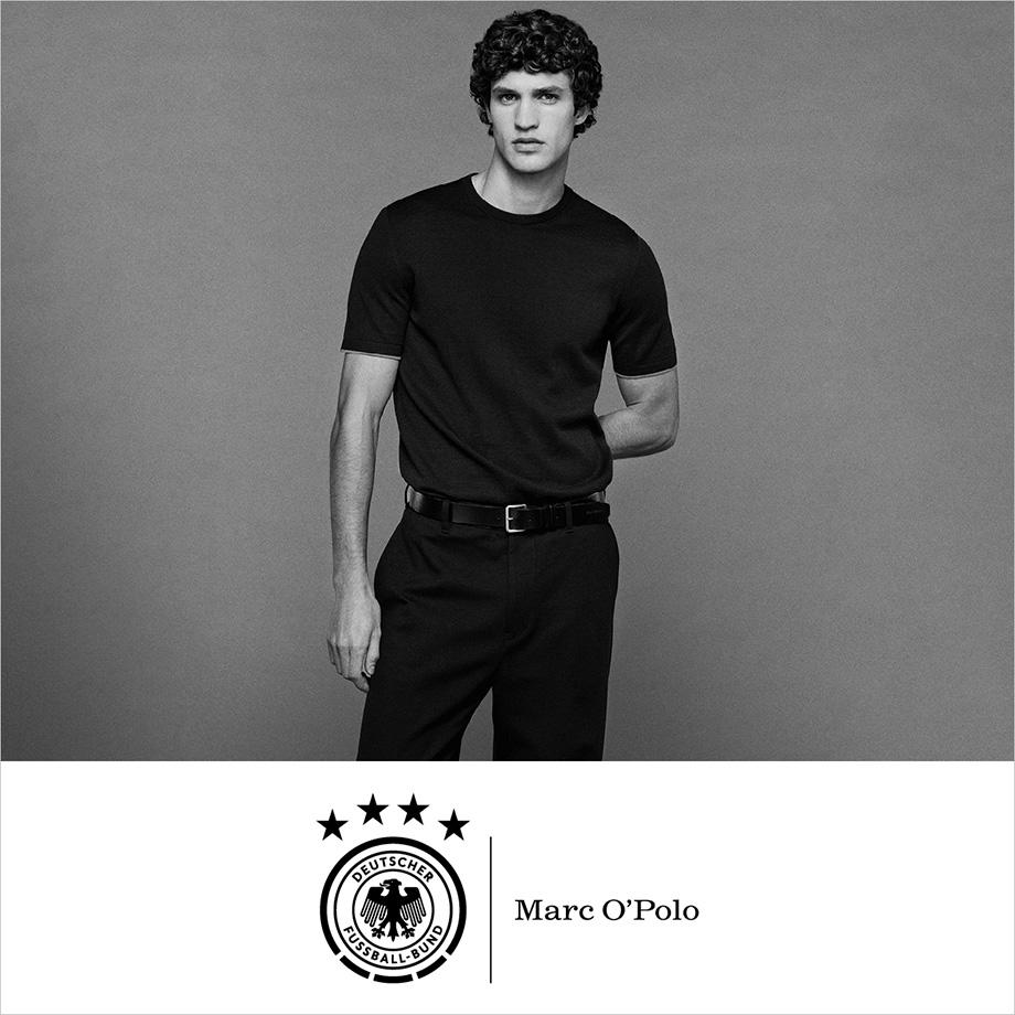 Schwarz-weiß Porträt eines Mannes in einem schwarzen Outfit von Marc O’Polo, Teil der Kooperation mit dem Deutschen Fußball-Bund; der Mann steht vor grauem Hintergrund.