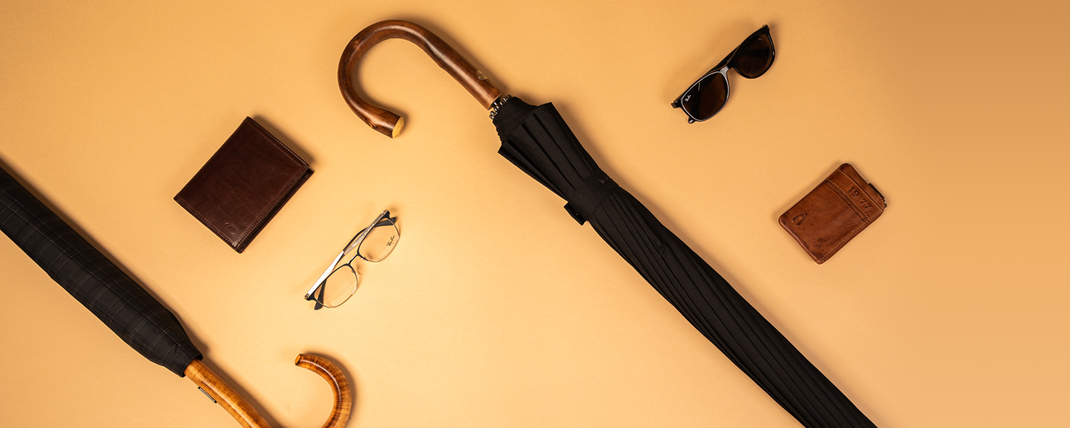 Schwarzer Regenschirm mit Holzgriff, Umgeben von braunem Lederportemonnaie, Sonnenbrille und Lesebrille auf beigem Hintergrund