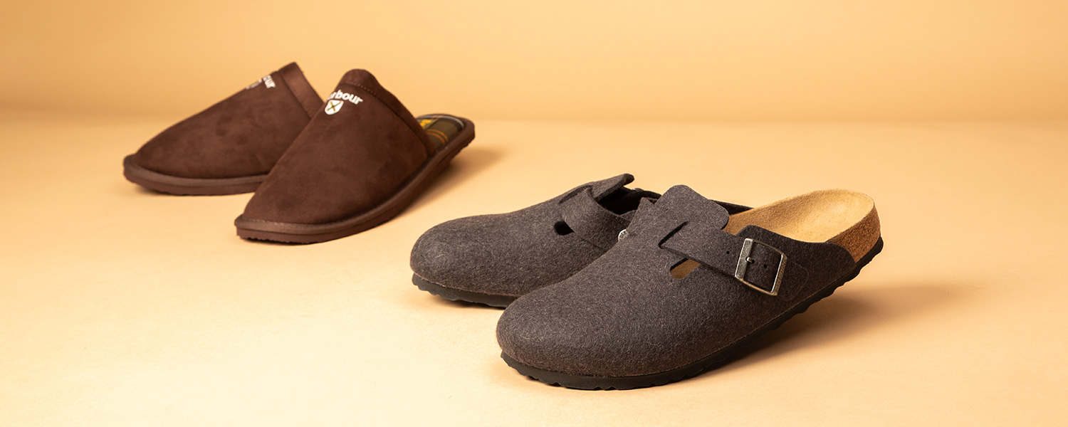 Graue Filz-Pantoffeln mit Schnalle und braune Hausschuhe aus Lederimitat auf beigem Hintergrund präsentiert.