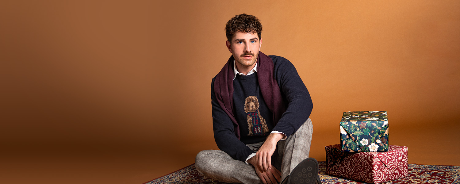 Herr in dunkelblauem Strickpullover mit Hunde-Motiv und grauer Hose auf Teppich sitzend