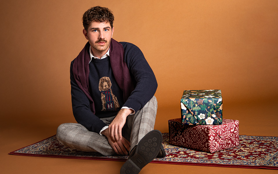 Mann in dunkelblauem Strickpullover mit Hundemotiv und karierten Hosen auf Teppich sitzend