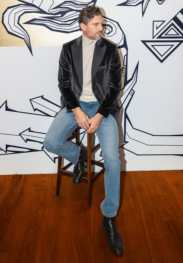 Herr in schwarzem Samtblazer mit Rollkragenpullover und blauen Jeans, sitzend auf einem Hocker