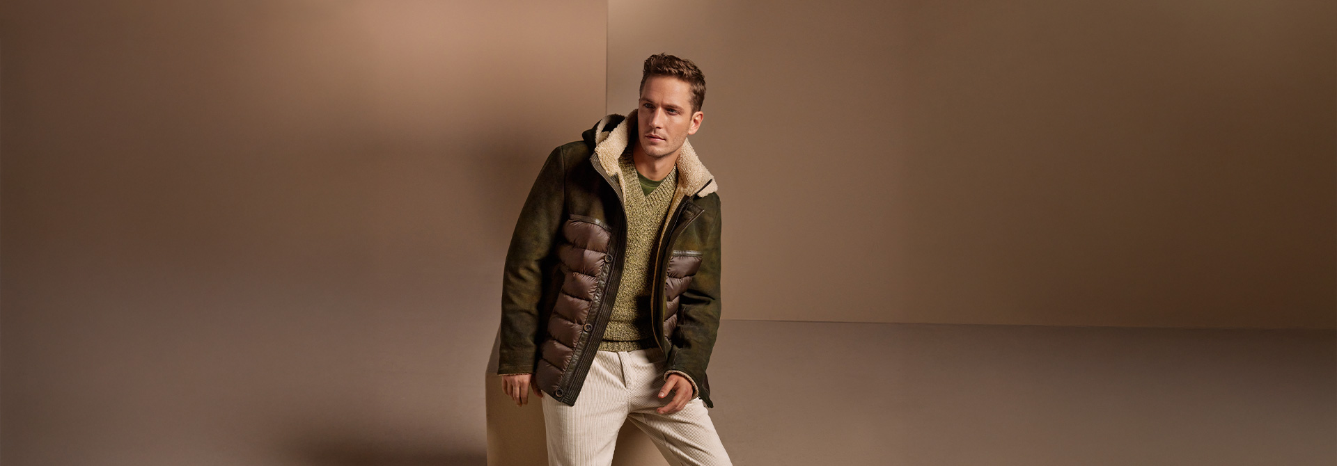 Mann in grüner Daunenjacke mit Schafsfellkragen und Strickpulli, beige Hose