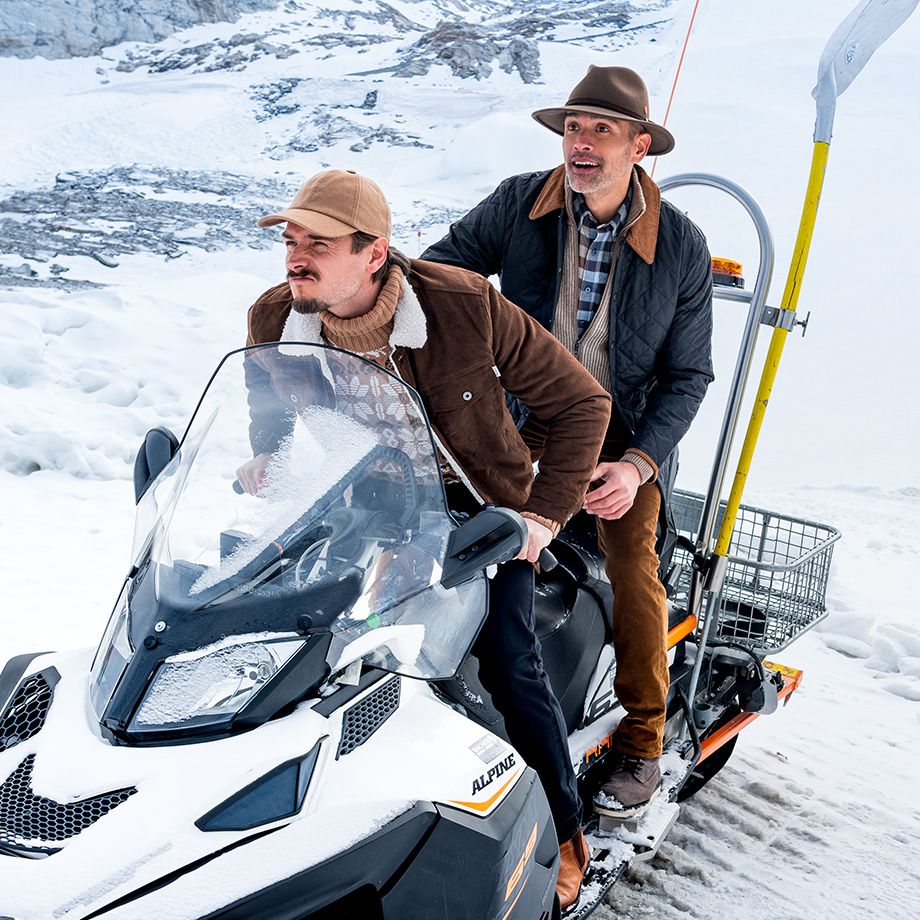 Zwei Männer auf Schneemobil, einer trägt braune Jacke mit Schal, der andere gesteppten Mantel, in winterlicher Landschaft