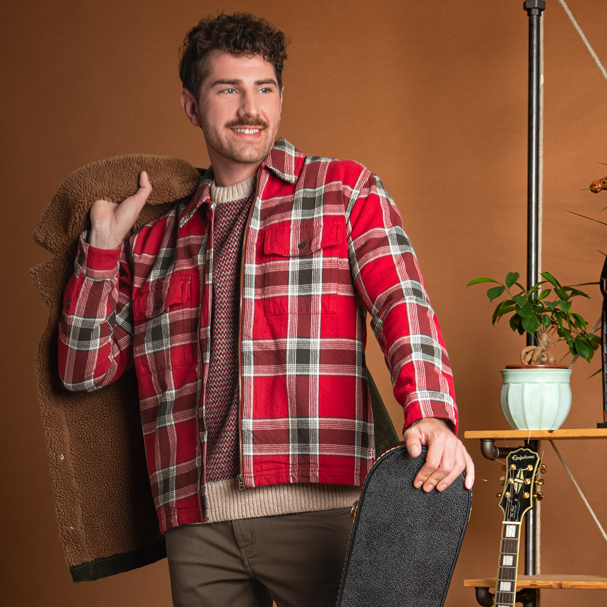 Mann in roter karierten Jacke aus Baumwolle, leger mit beigem Pullover getragen, hält Gitarrenkoffer
