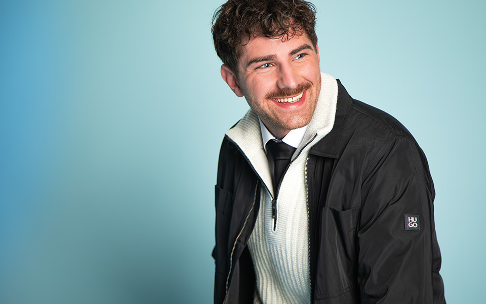 Mann in schwarzer Jacke über weißem Pullunder und grauer Anzughose, lässig sitzend