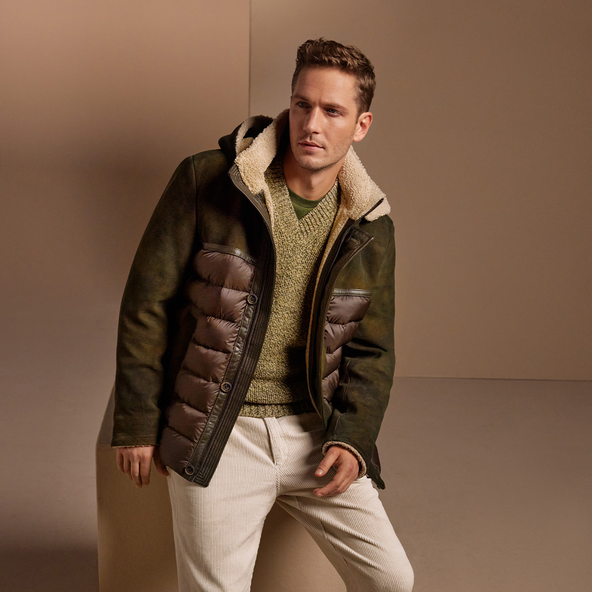 Mann in olivgrüner Übergangsjacke mit Kapuze, darüber grauer Strickpullover, beige Cordhose