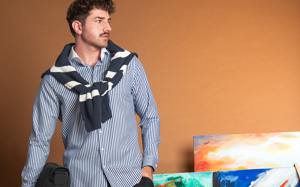 Mann in blau-weiß gestreiftem Freizeithemd mit Pullover über den Schultern vor Kunstwerken