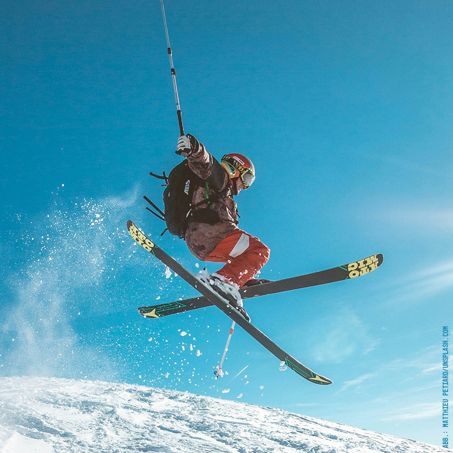 Skifahrer in roter Hose und brauner Jacke beim Sprung vor klarem Himmel