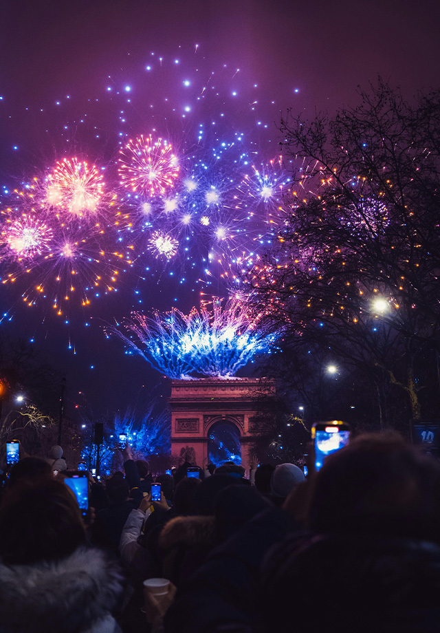 Farbenprächtiges Feuerwerk über Torbogen bei Nacht, beobachtet von einer Menschenmenge, die das Spektakel mit Smartphones fotografiert.