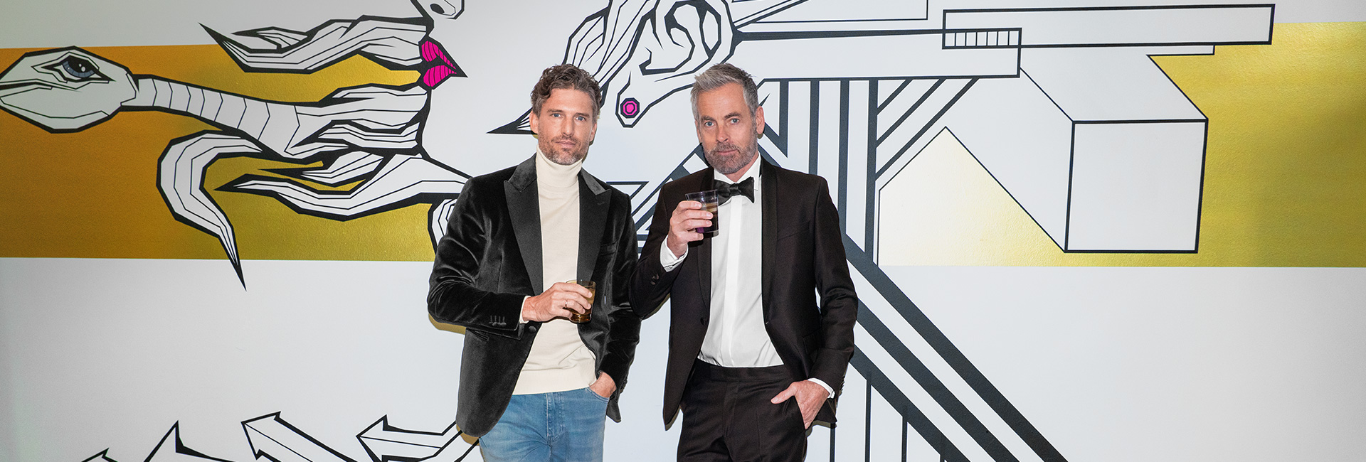 Herren im schwarzen Smoking und schwarzem Samtblazer mit Drinks, vor grafischem Hintergrund stehend.