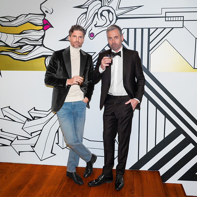 Herren im schwarzen Smoking und schwarzem Samtblazer mit Drinks, vor grafischem Hintergrund stehend.