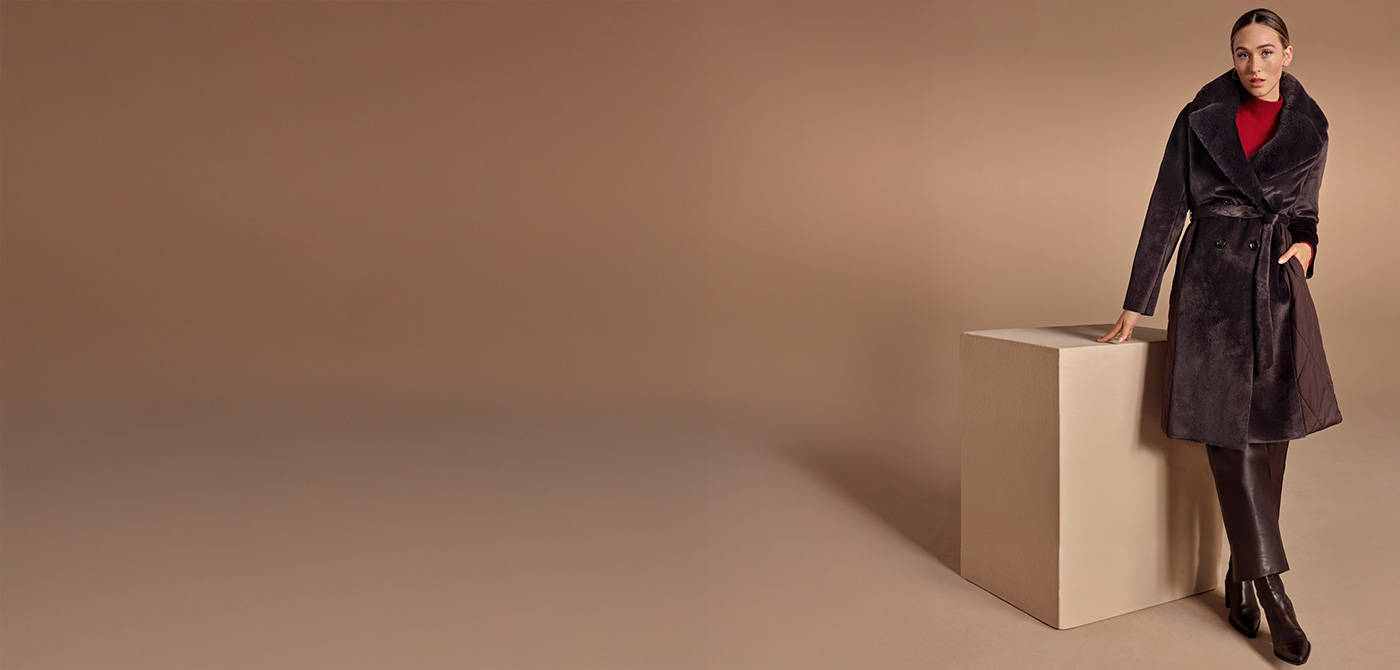 Frau in dunkelbraunem Mantel mit Gürtel und rotem Pullover, gegen beigen Hintergrund