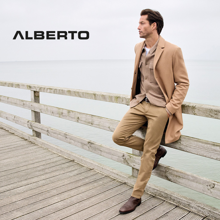 Mann in beige Chinohose mit beigem Mantel an der Küste, elegant kombiniert